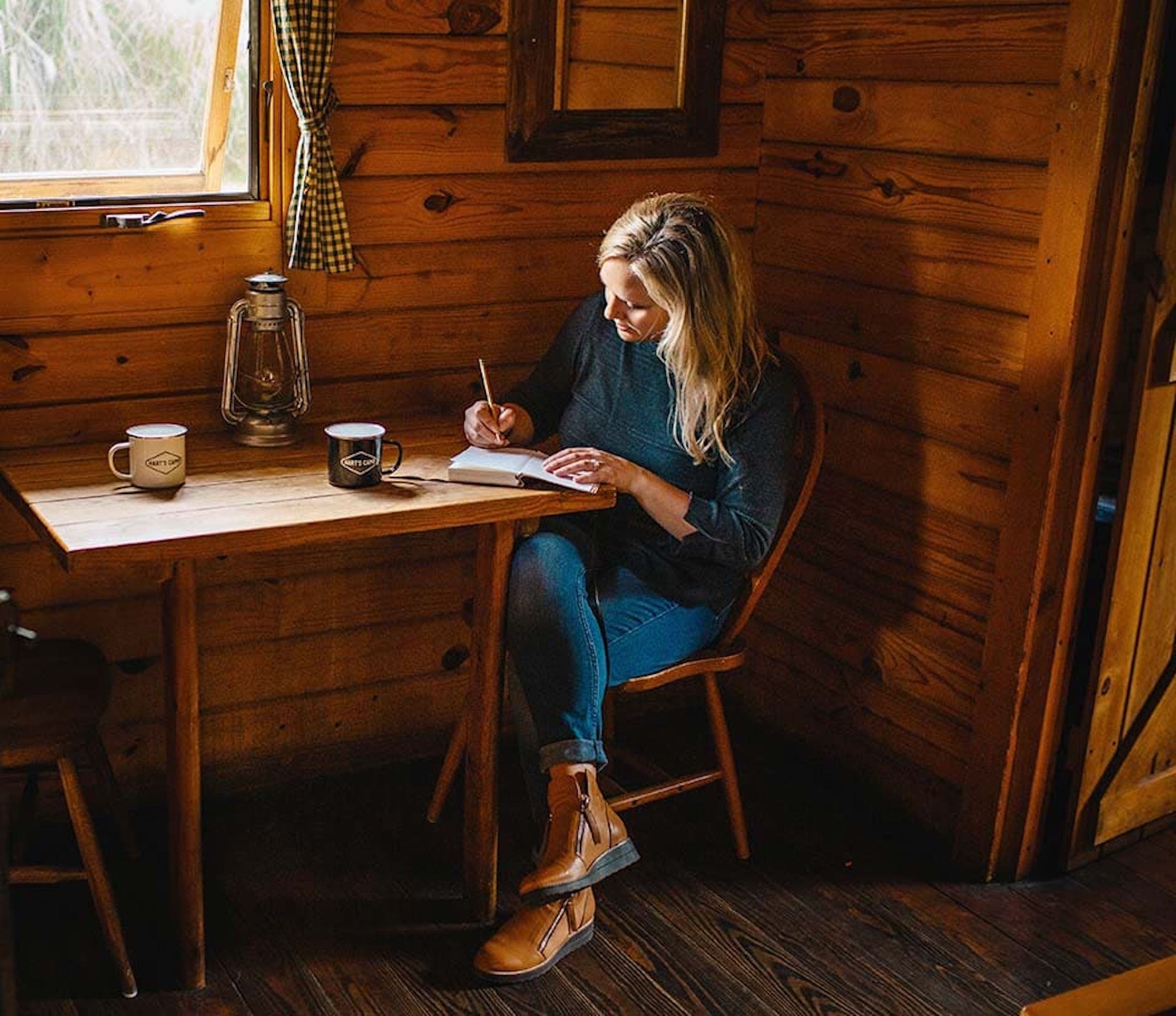 A woman sitting at a wooden table while writing something in a book in a cabin at Harts Camp
